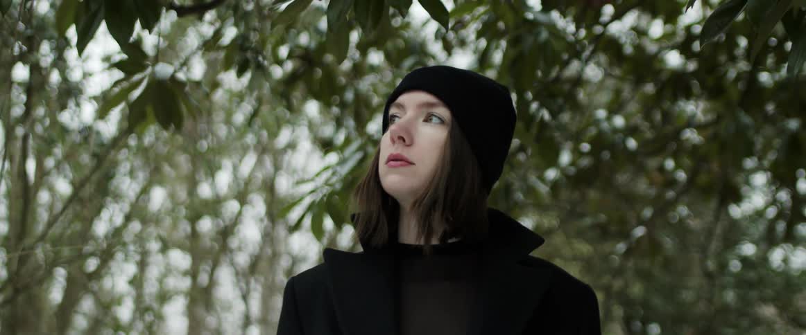 Woman wearing a black knit hat and a black coat is walking among trees and looking around