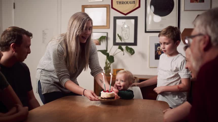 A mom carries a cake with two tall candles to the table and places in front of a little girl as family looks on