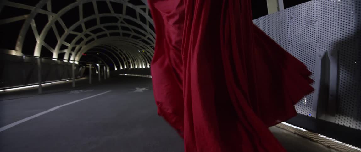 A woman with a red dress walking in an overpass with buildings and the sky in the background