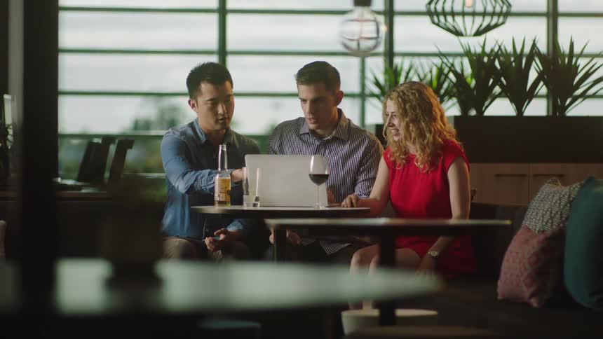 Group of friends sitting at a table in a restaurant looking at a laptop screen while chatting and laughing