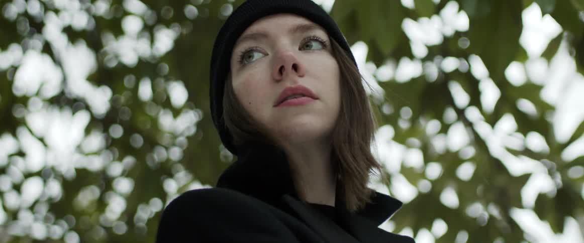 Woman wearing a black knit hat and a black coat is looking around with trees behind her