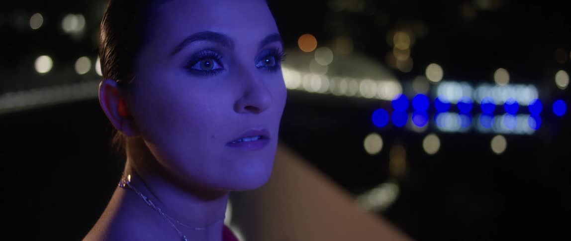 A woman with a necklace and a red dress on an overpass with lights in the background