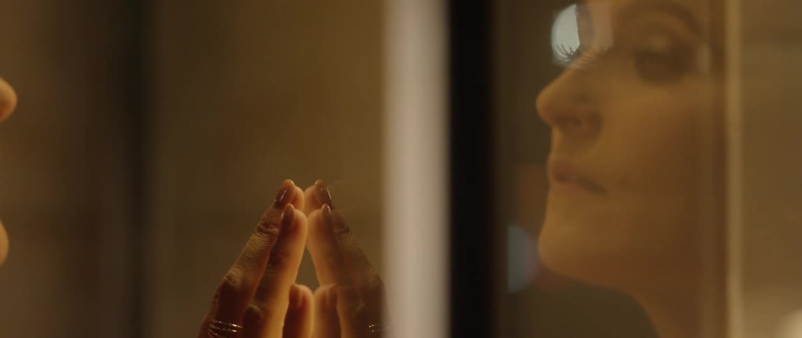 A woman puts her manicured hand against a wall and looks out a window