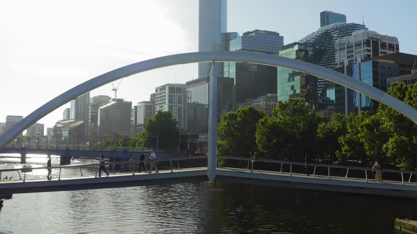 People walking on a bridge over a river with buildings skyscrapers the sky and clouds in the background