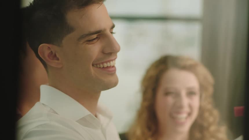 A young man and woman laugh as the man writes on a whiteboard and then another man comes up and pats him on the back