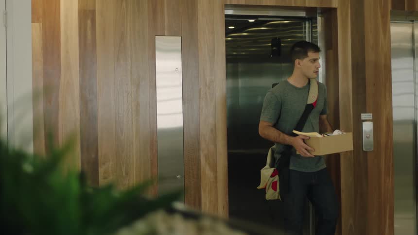 A man with a backpack and a box walking out of an elevator with plants in the foreground