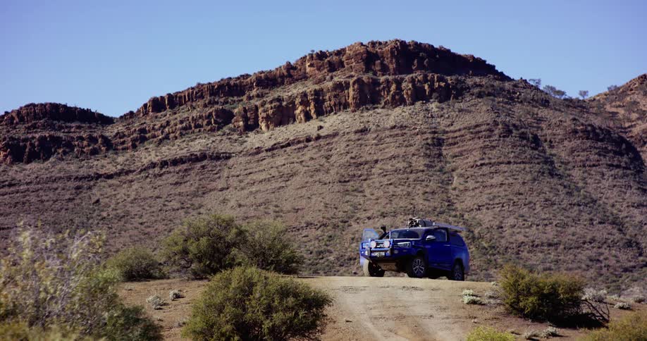 Two people getting in and out of a blue truck parked in front of a mesa plateau on a clear day