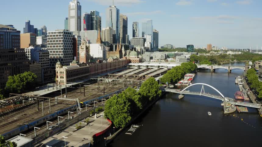 An aerial view of downtown area with a train driving alongside a river and people walking on a bridge and a boardwalk