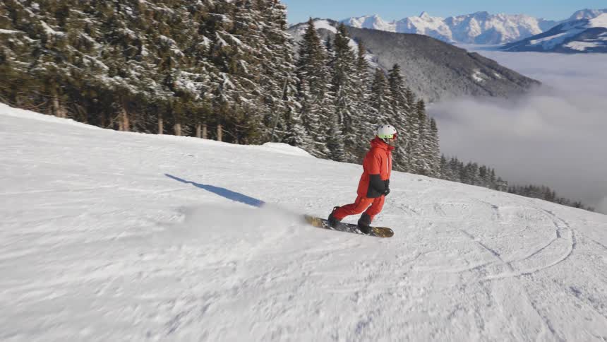 Person wearing an orange snow suit and white helmet is riding a snowboard down a hill with many pine trees and mountains around