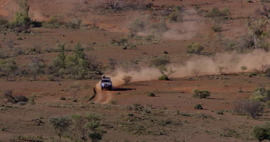 A vehicle driving on a dirt road raisin dust with shrubs and trees in the background