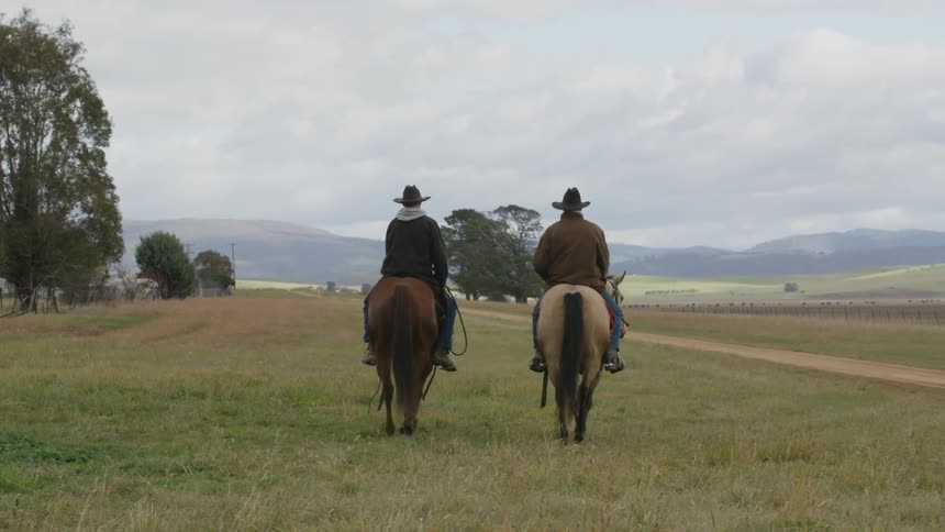 Two men riding horses with cowboy hats with trees the sky and clouds in the background