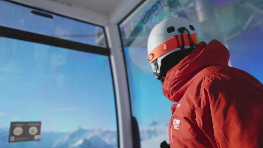 A person sitting in a ski lift points out the window towards the distant snow covered mountain