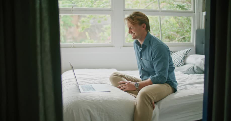 Man with blue shirt and brown pants sits on bed and laughs while making video call on laptop in bedroom
