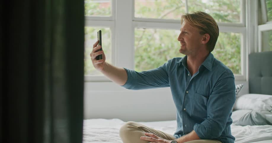 A man in a bedroom sitting on a bed video chatting on a smart phone