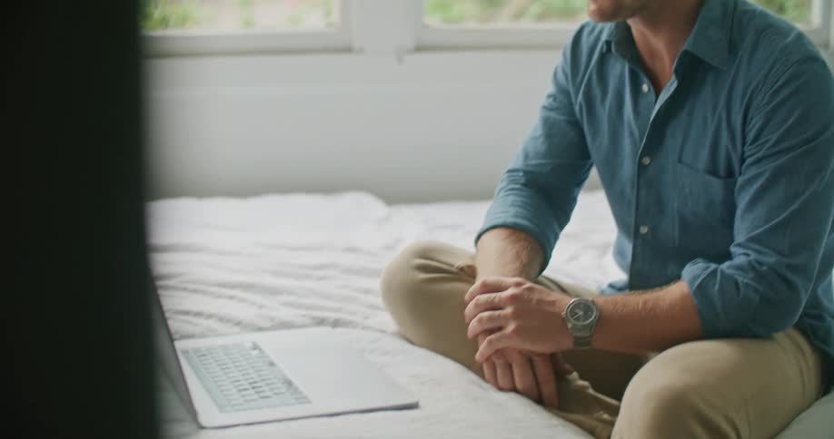 A middle aged man sitting on a bed laughs while using a laptop to participate in a video chat