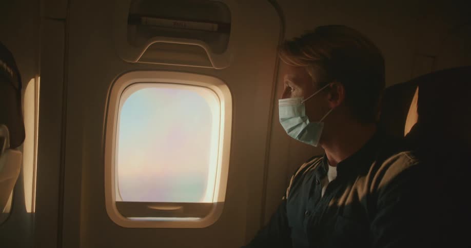 A man with a mask in an airplane sitting on a chair looking out of the window