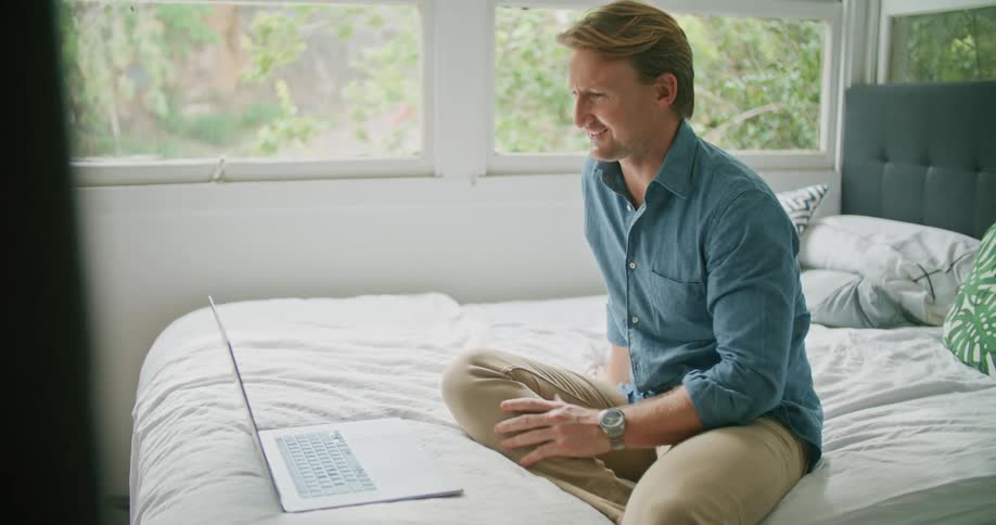 A man in a bedroom sitting on a bed video chatting on a laptop with windows and a wall in the background