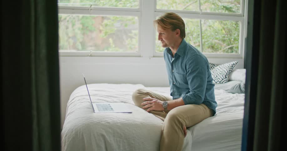 A man sits on his bed at home and talks with someone on his laptop