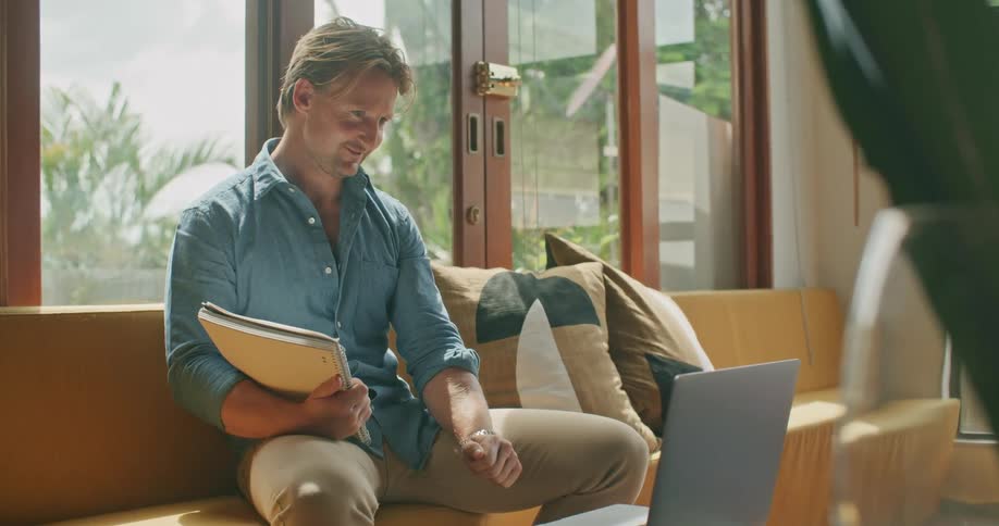 White man sitting at a couch on a video call while holding a book