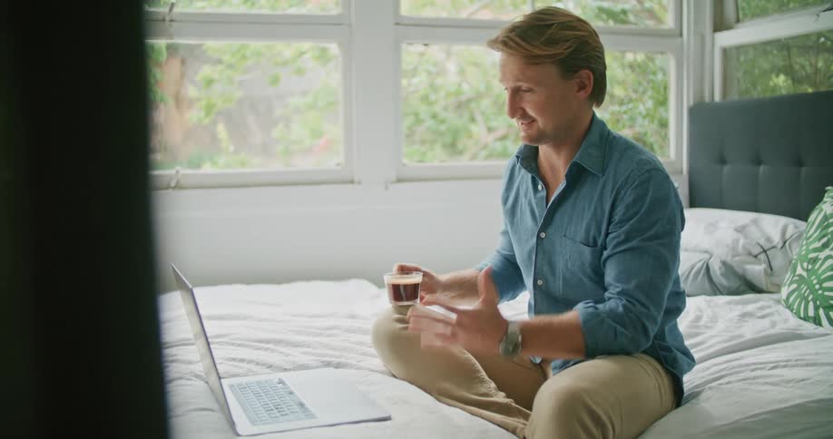 A man drinks coffee while sitting in bed and talking to someone on his laptop