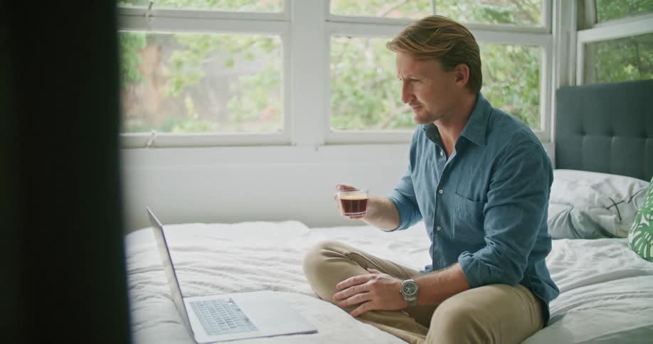 Man sitting on bed drinking coffee blows kiss to person on other end of video call