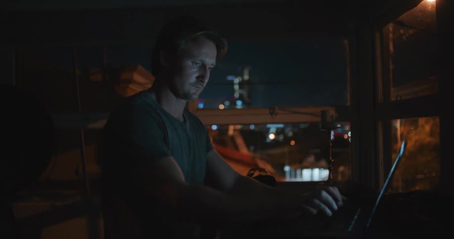 A man types and works at a laptop near large windows at night
