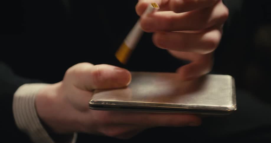 A man removes a cigarette from a metal case and taps the cigarette on it  after closing