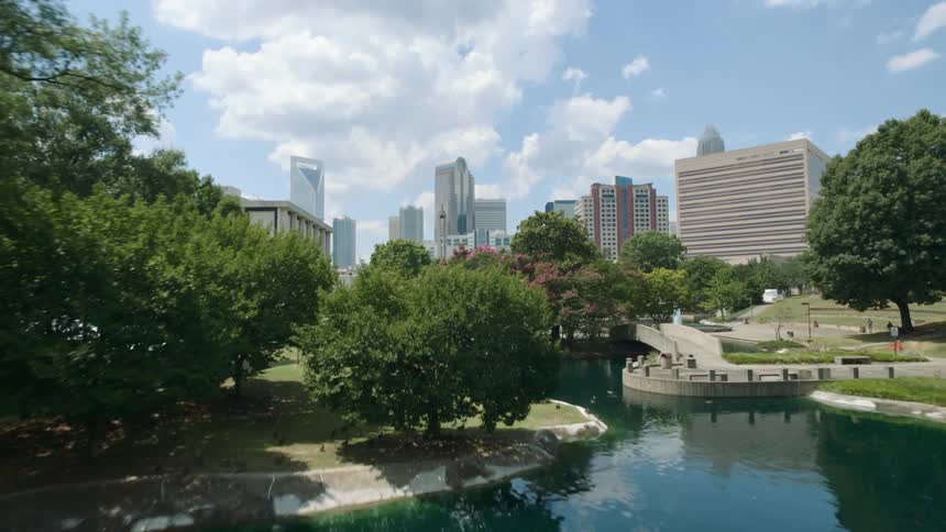 A city skyline stands behind a park with a shimmering pond on a bright blue sky day