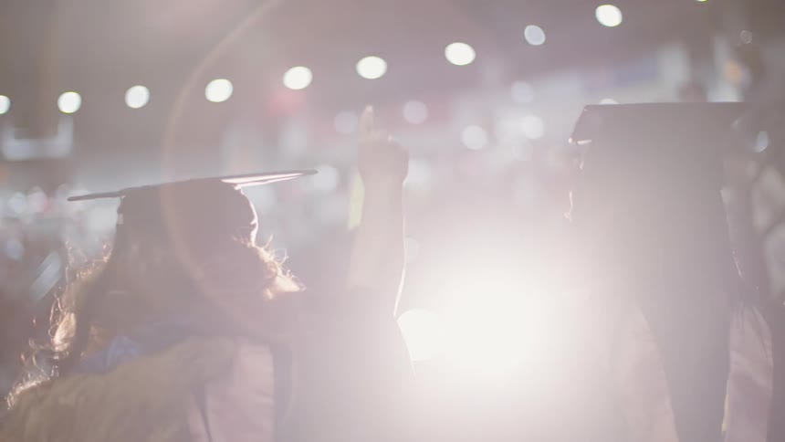 A graduating student raises her hand as she and two friends enter an auditorium