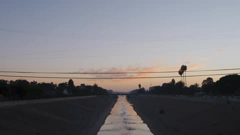 A canal with water with trees an overpass vehicles the sky and clouds in the background
