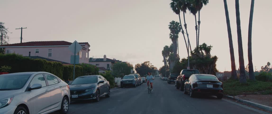 A woman rides her bicycle down the middle of a neighborhood street lined with parked cars