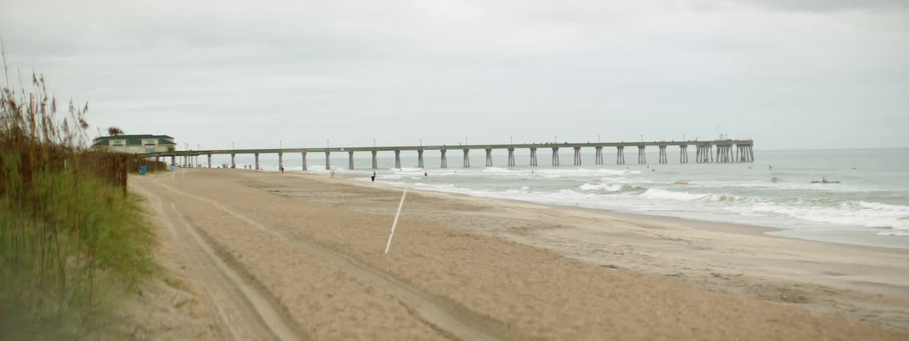 A long pier juts out into the ocean as people surf and boat