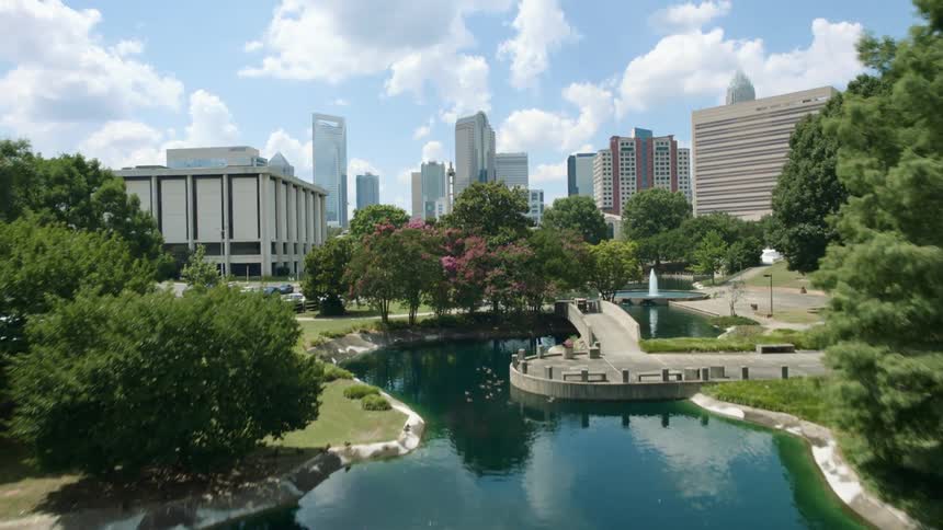The pond water surface shimmers and reflects the blue sky and clouds with city buildings behind the park