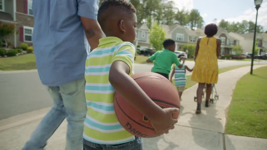 A boy with a basketball walking on a sidewalk holding a mans hand with two boys and a woman pushing a stroller in front with houses the sky and clouds in the background