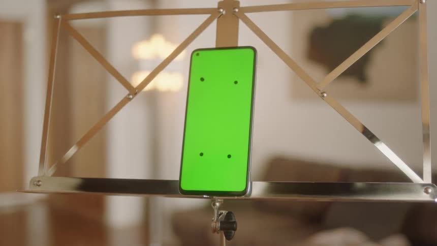 Cinematic circular tracking shot around smartphone with green screen and tracking marks mounted on gold metal music stand in bright living room. Filmed in soft natural light, perfect for VFX, app mockups, and digital product presentations.