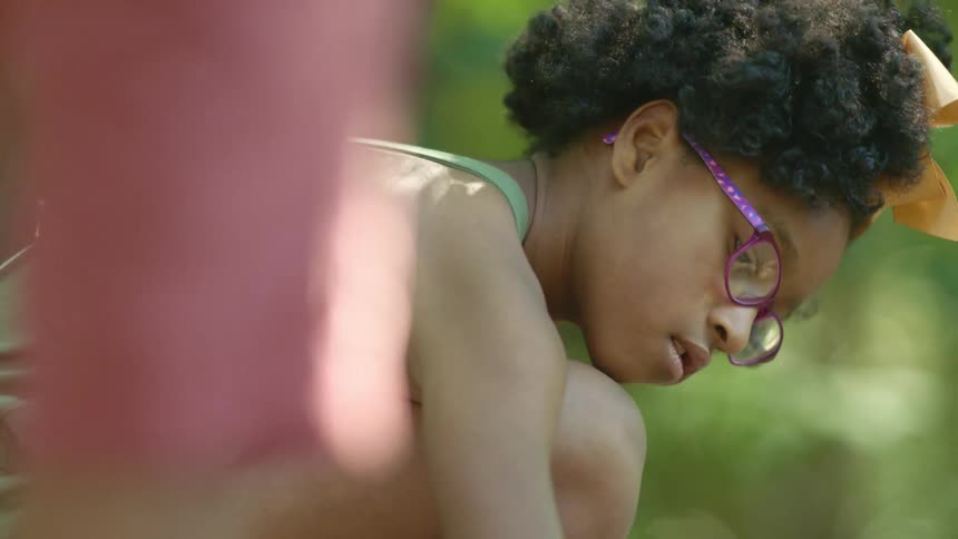 A little girl with glasses and a bow in her hair stooping down with trees in the background
