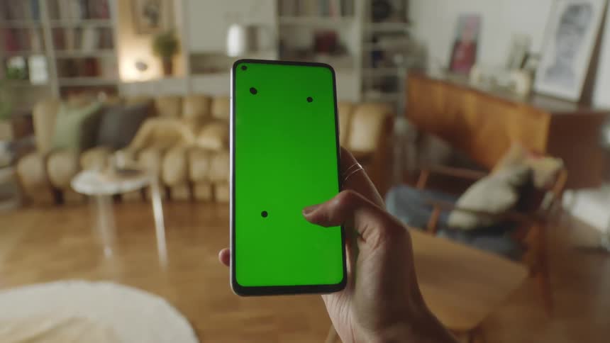 Person walking through cozy living room holding smartphone with green screen and tracking marks, tapping screen before lowering it out of frame. Filmed in natural light, perfect for VFX, app mockups, and digital lifestyle projects.