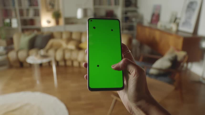 Person walking through cozy living room holding smartphone with green screen and tracking marks, tapping screen before lowering it out of frame. Filmed in natural light, perfect for VFX, app mockups, and digital lifestyle projects.