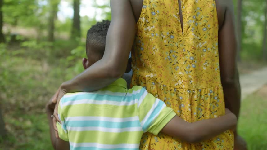 A boy embraces his mother as they walk along a paved walkway through a woodland during the day