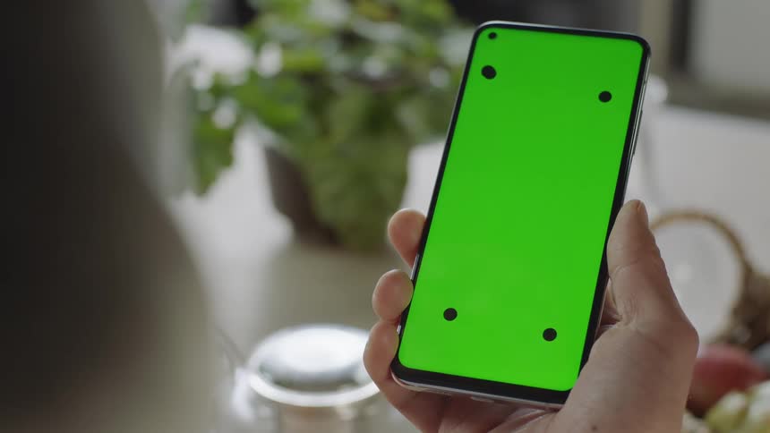 Close-up of a hand holding a smartphone with green screen and tracking marks, filmed indoors in soft natural light. Hand moves device out of frame for a smooth transition. Ideal for VFX, screen replacement, and app mockup projects.