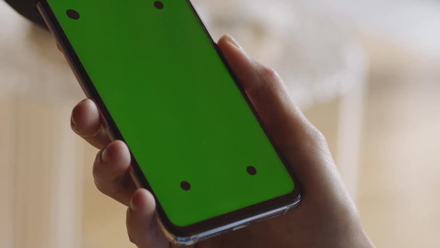 A detailed close-up shot of a hand holding a smartphone with a green screen and tracking markers. Filmed indoors in soft daylight, the static shot provides a clean and versatile VFX plate for screen replacement, app mockups, or post-production projects.