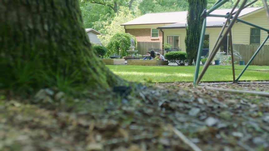 A woman instructs her children as they kneel before a planter in the yard of a house during the day