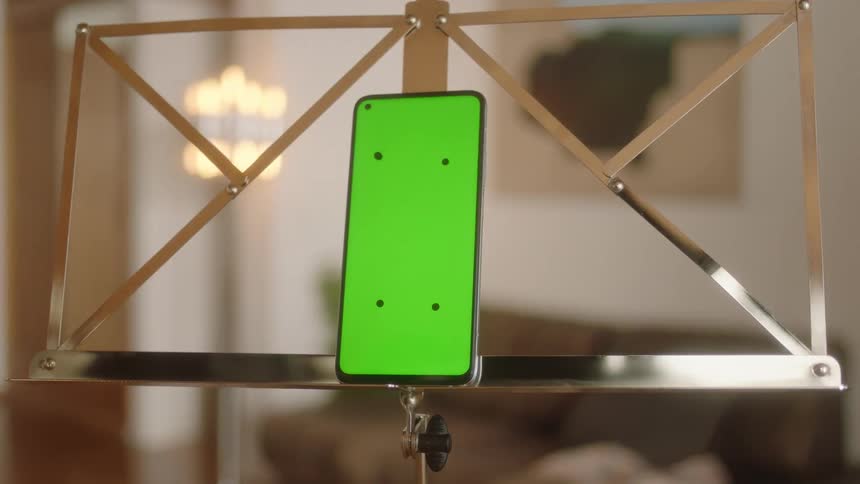 Smooth slow circular camera move around smartphone with green screen and tracking marks placed on elegant gold metal music stand in cozy living room. Filmed in soft daylight, ideal for VFX, app mockups, and technology presentation projects.