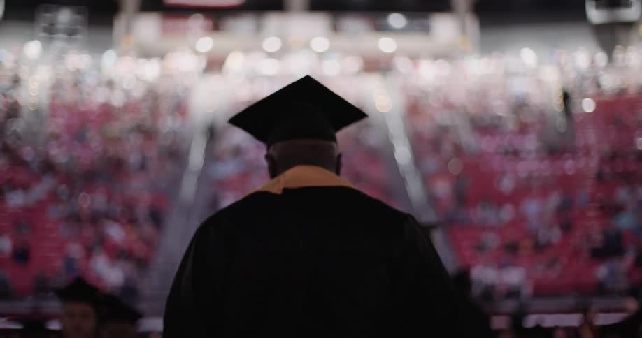 A man wearing a graduation cap and gown walks in an auditorium