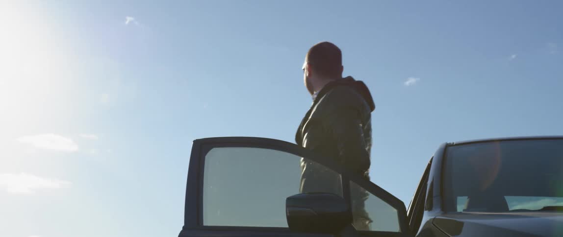 A man exits a car parked on the side of a dirt road on a sunny day