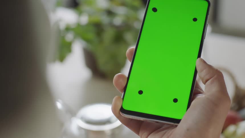 A close-up of a hand holding a smartphone with a green screen and tracking markers, filmed indoors in soft natural light. At the end of the shot, the hand moves the device out of frame. Perfect for visual effects, screen replacement and app mockups.