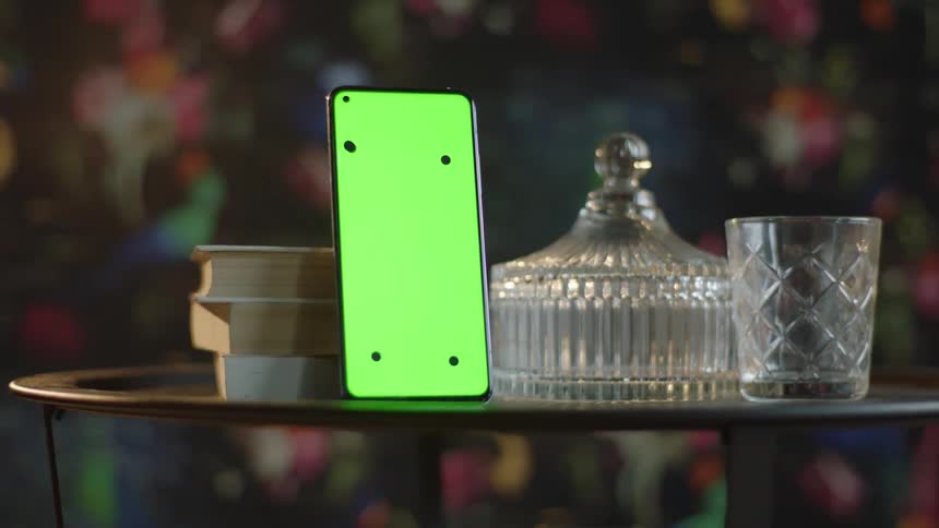 Cinematic Circular Tracking Shot Around Smartphone with Green Screen and Tracking Marks on Table with Glassware and Books. Indoor Scene for VFX and App Mockups.