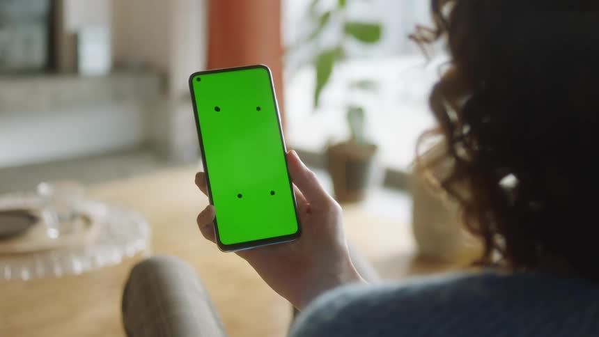 Woman with curly hair sitting in bright living room raises smartphone with green screen and tracking marks to look at it, then lowers it again. Filmed in soft daylight, perfect for VFX, app mockups, and digital lifestyle projects.