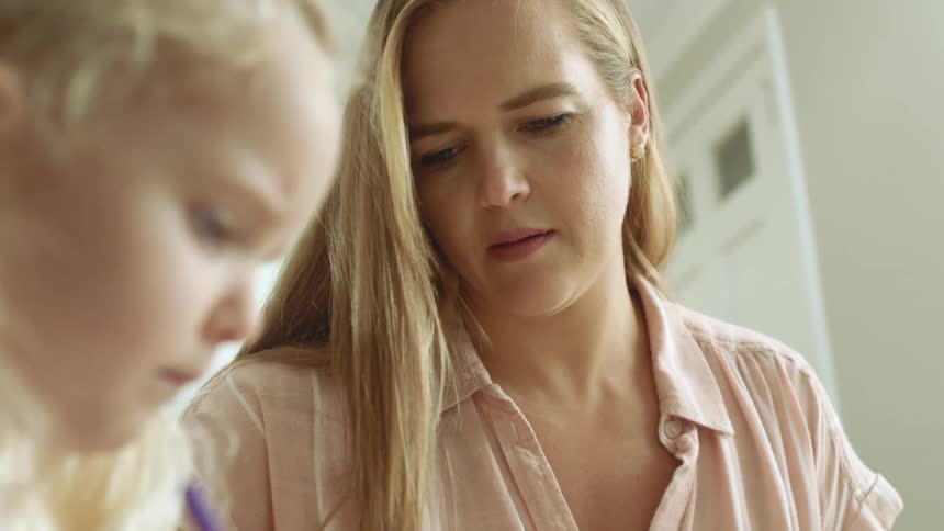 A woman sitting next to her daughter at a table instructs the girl as she writes