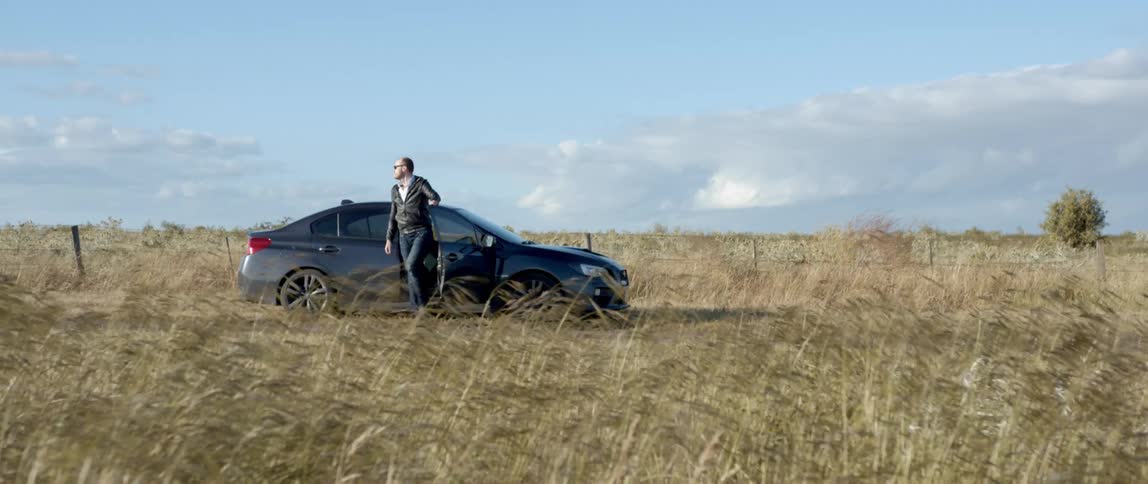 A man gets out of the passenger seat of a dark blue sedan parked in a field on a windy day